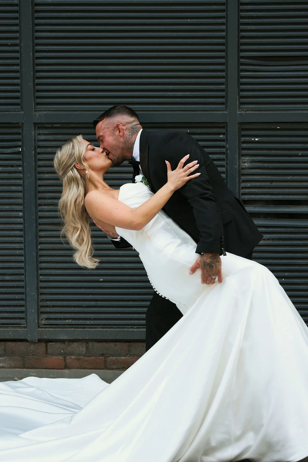 A newlywed couple shares a dramatic dip kiss against an urban slatted metal and brick backdrop, with the bride wearing a sleek strapless white satin gown.
