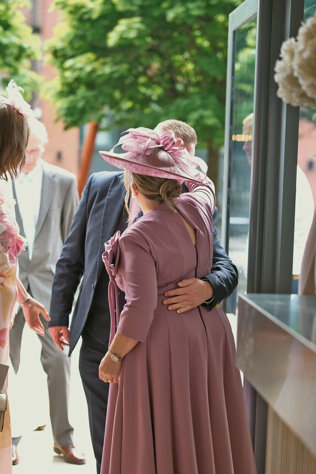 Wedding guests wearing a mauve dress with a matching fascinator and a grey suit greet each other indoors near a glass doorway.