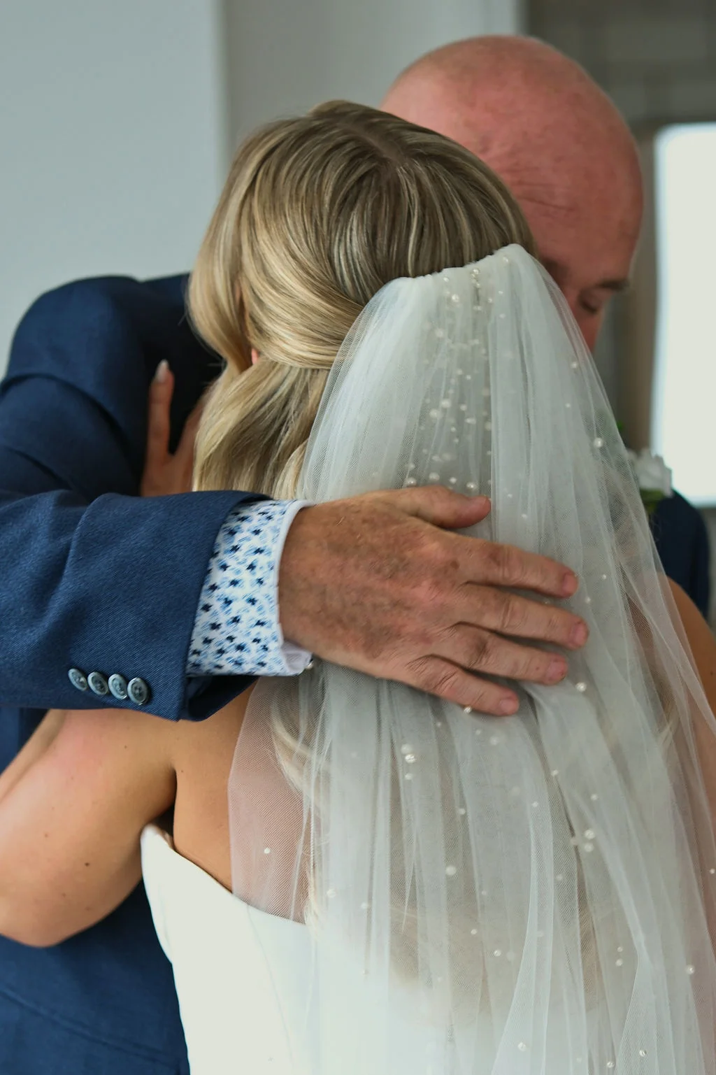 A close up back view of a father embracing his daughter in a wedding dress wearing a pearl veil and a navy suit.