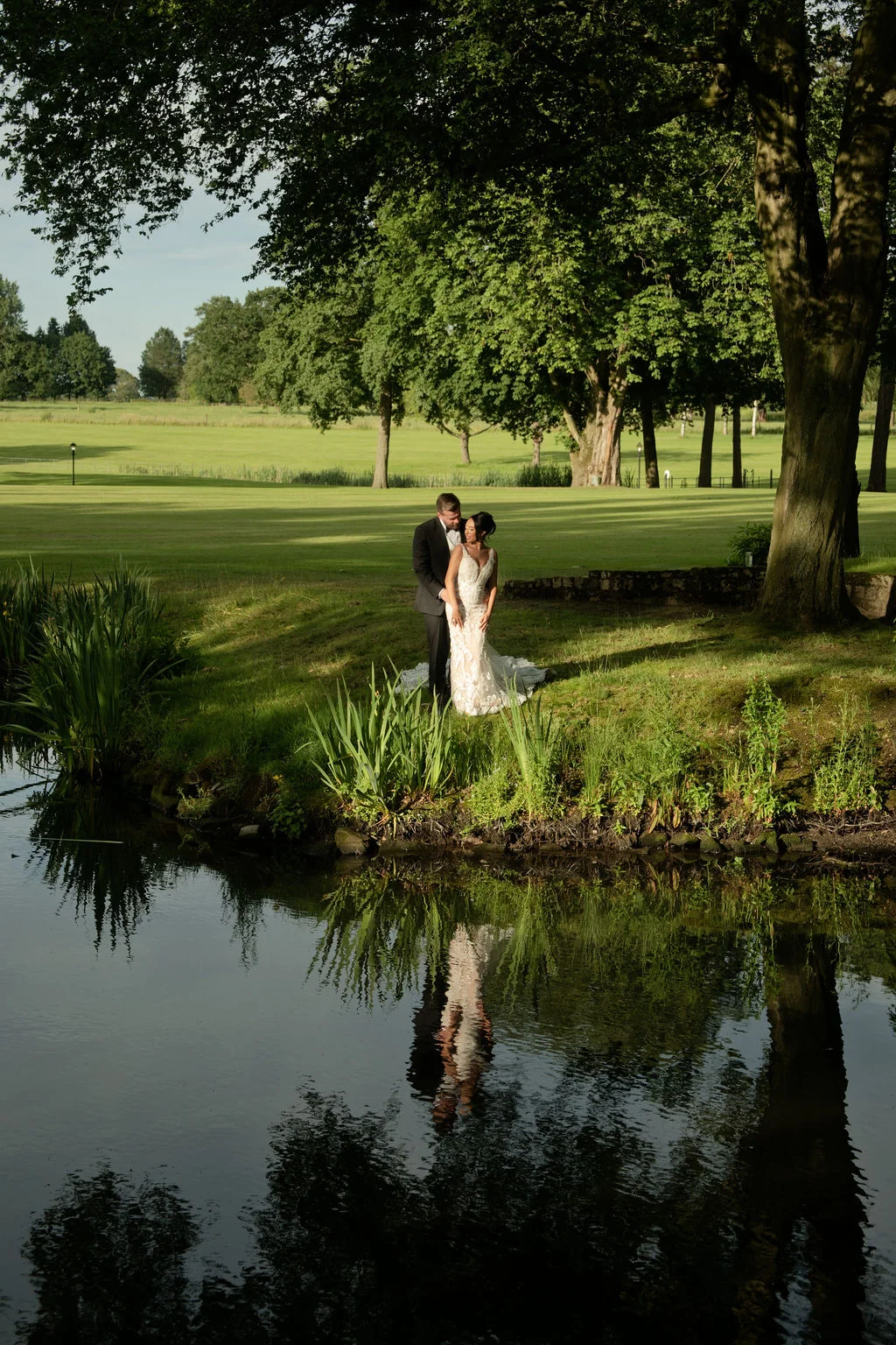 A newlywed couple stands embraced by a tranquil lake and mature trees during golden hour, casting a clear reflection in the water below.