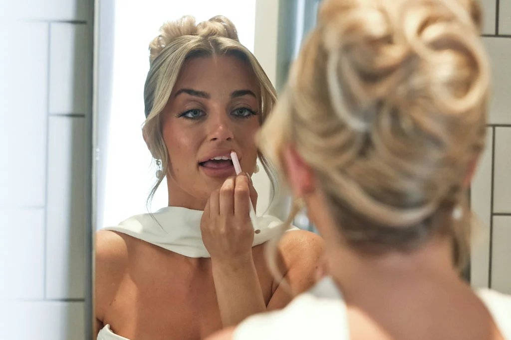 A blonde bride applies lipstick while looking into a mirror during her wedding morning preparations.