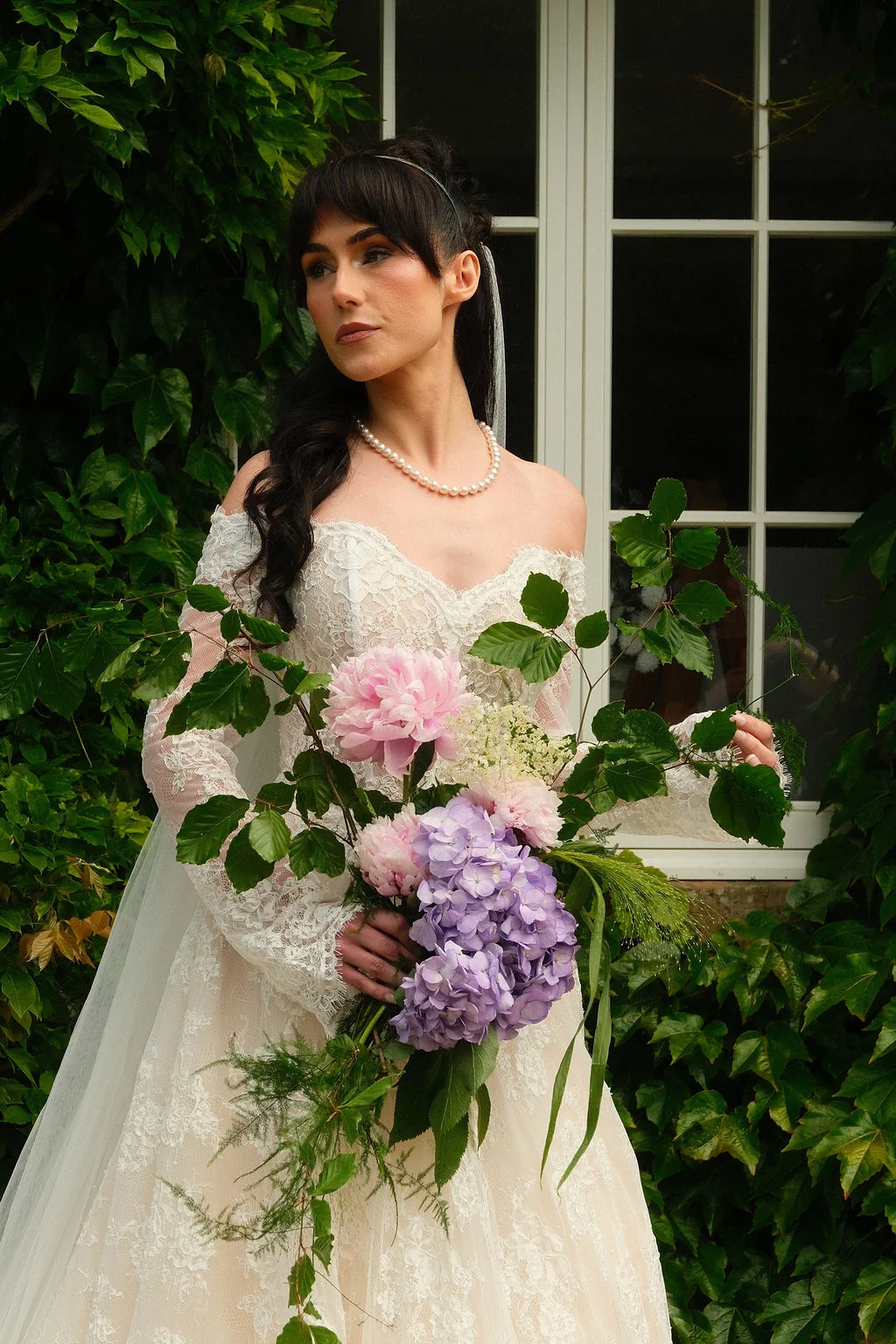 A bride with dark hair and a fringe poses against an ivy covered wall, wearing an off-the-shoulder lace gown and pearl necklace while holding an abundant trailing bouquet of peonies and hydrangeas.
