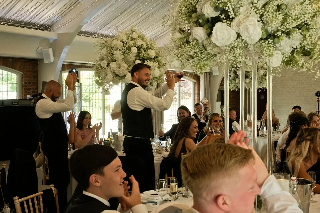 A groom in a waistcoat raises a glass during the wedding speeches as guests cheer and applaud in a brightly lit barn venue.