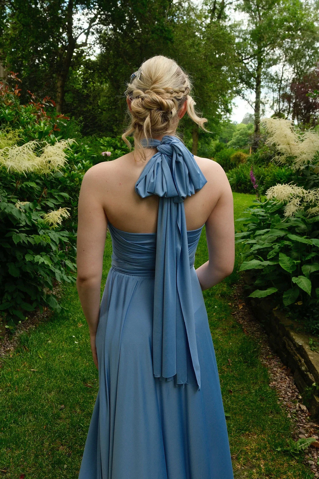 A bridesmaid stands in a lush garden wearing a dusty blue multi-way gown featuring a halterneck and an elegant knotted bow at the back, paired with a braided updo.