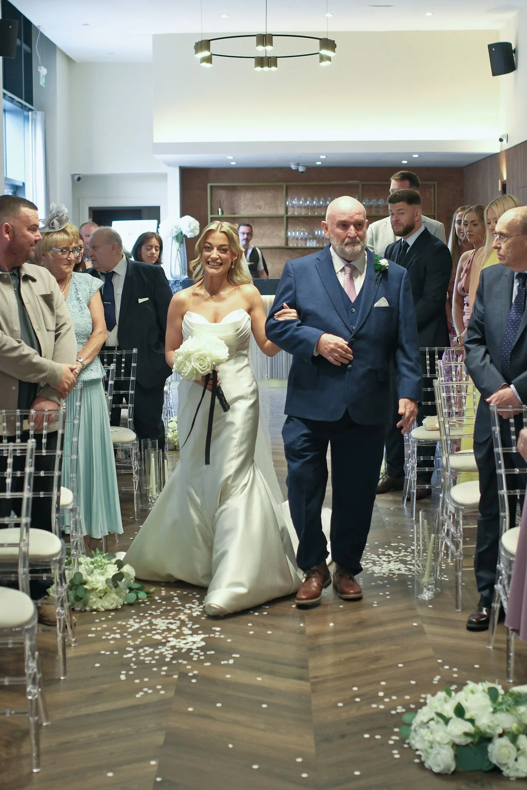 A smiling bride holding a bouquet of white roses walks down the aisle arm in arm with her father during a modern hotel wedding ceremony.