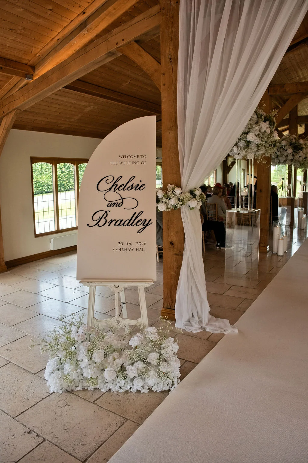 An elegant modern wedding welcome sign featuring custom black typography on a half-arch board is displayed on a white easel, surrounded by white floral arrangements inside a rustic oak-framed barn venue.