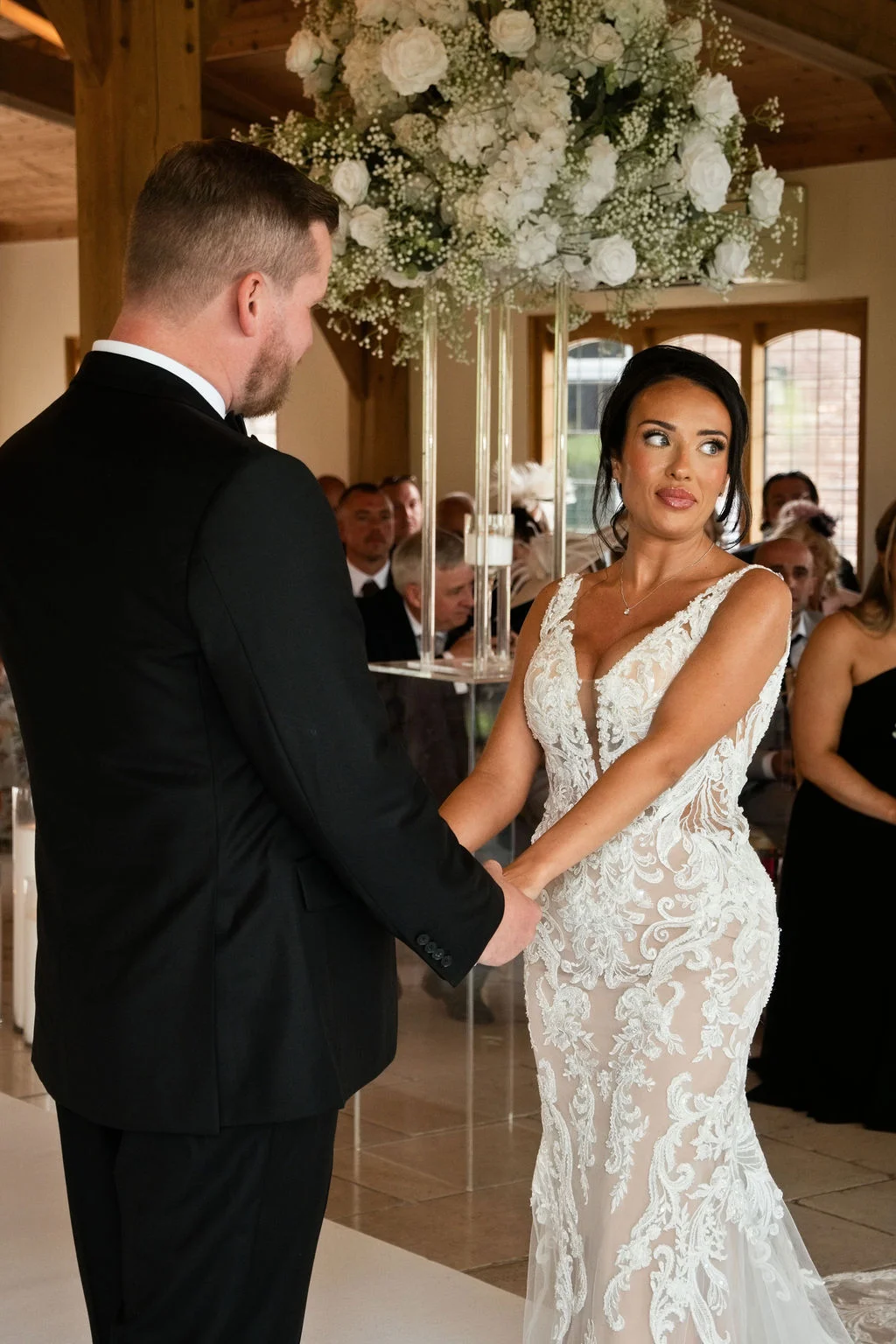A bride and groom holding hands during their wedding ceremony in a rustic barn venue with a large white floral hanging installation above.