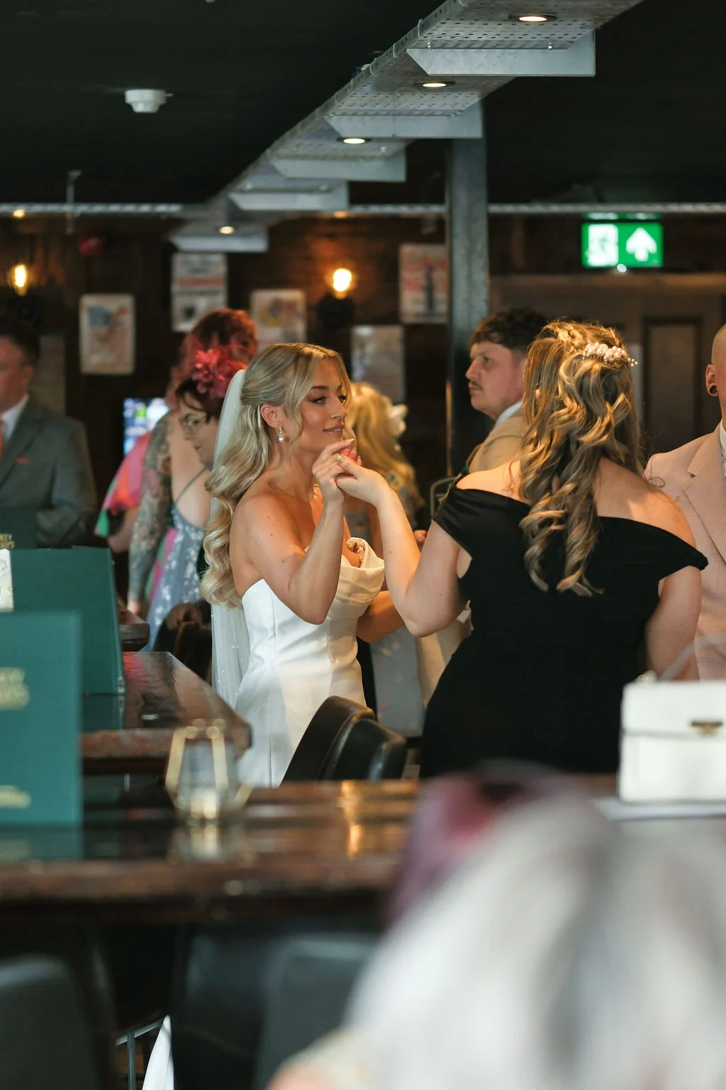A blonde bride wearing a strapless satin gown and holding hands with a guest inside a rustic pub venue with exposed beams.