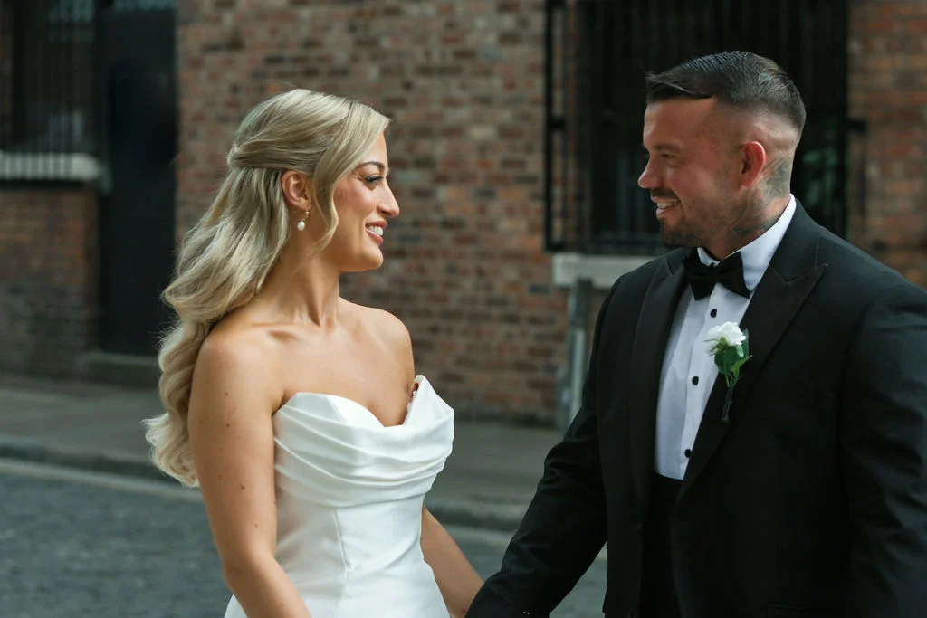 A newlywed bride and groom exchange a loving glance outside against an exposed brick wall, with the bride wearing a strapless white gown and the groom in a classic black tuxedo.
