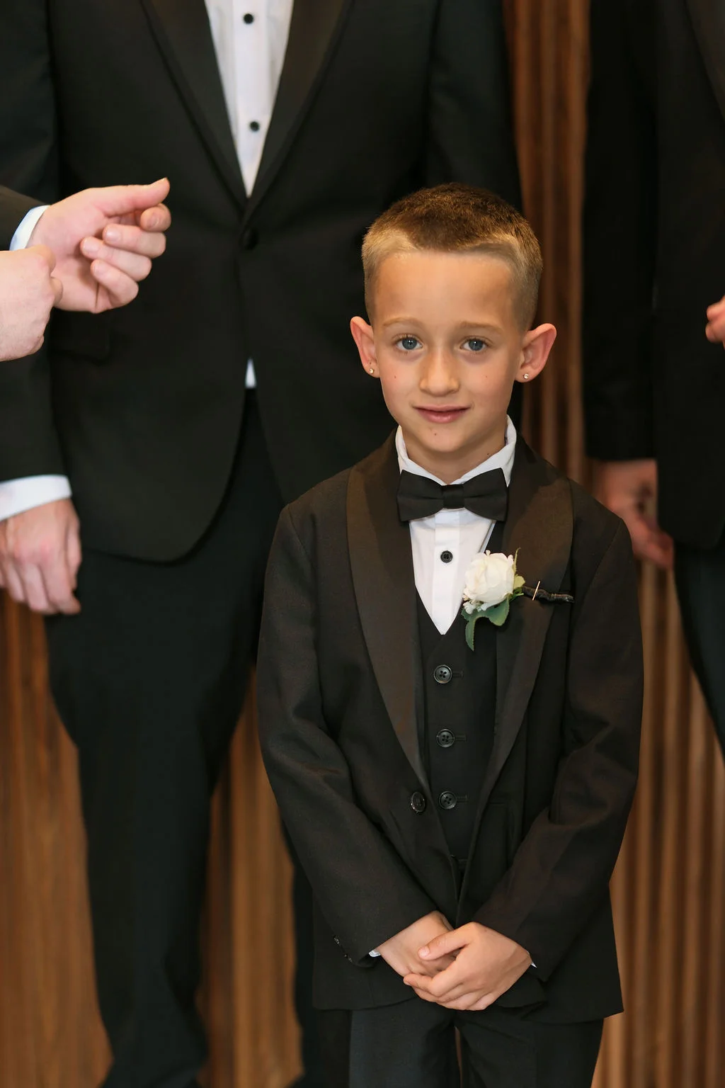 A young page boy wearing a smart black tuxedo with a matching bow tie and a white rose buttonhole stands among groomsmen.