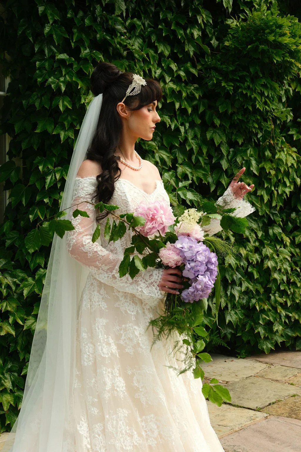 A bride with dark hair styled in an updo and crystal hairpiece stands in profile against an ivy-covered wall, wearing a long-sleeved lace wedding dress and holding a cascading bouquet of pink and purple flowers.