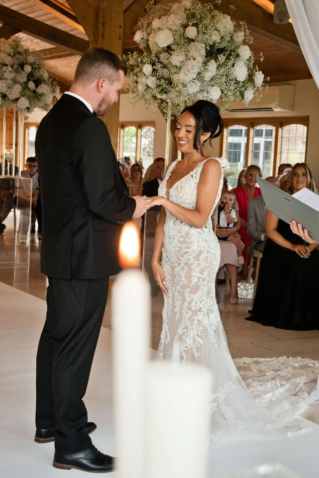 A groom slips a ring onto his bride's finger during a heartfelt ceremony inside a bright barn venue adorned with white floral arrangements.