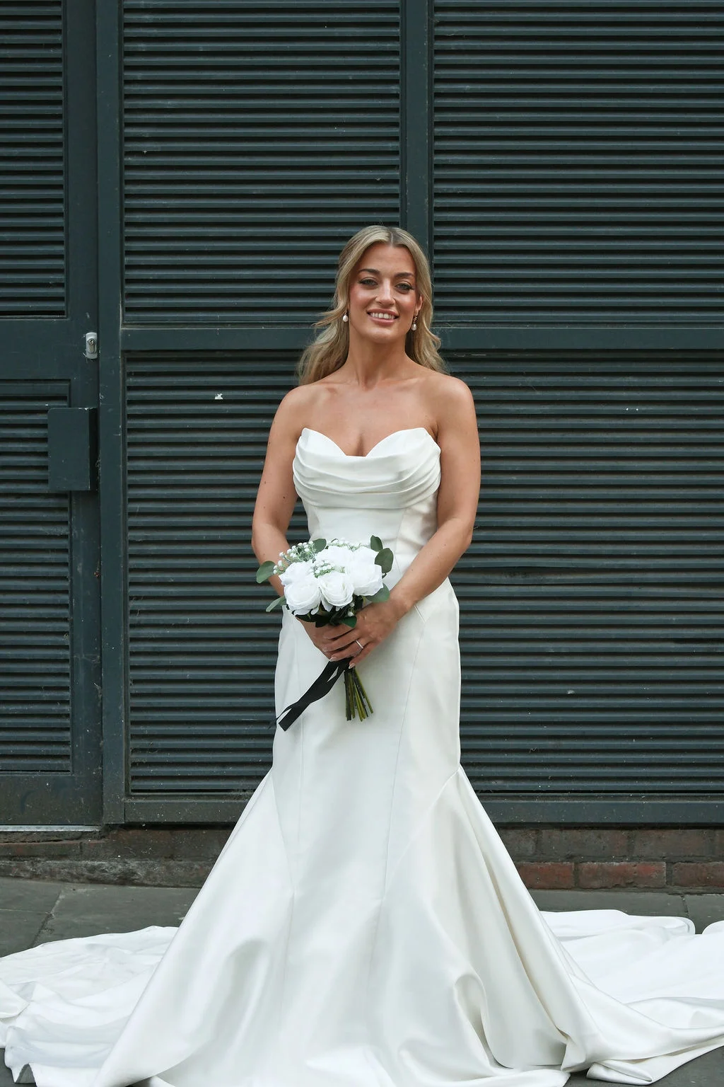 A stunning bride stands smiling in a sleek strapless white satin mermaid gown, holding a hand-tied bouquet of white roses against a dark industrial backdrop.