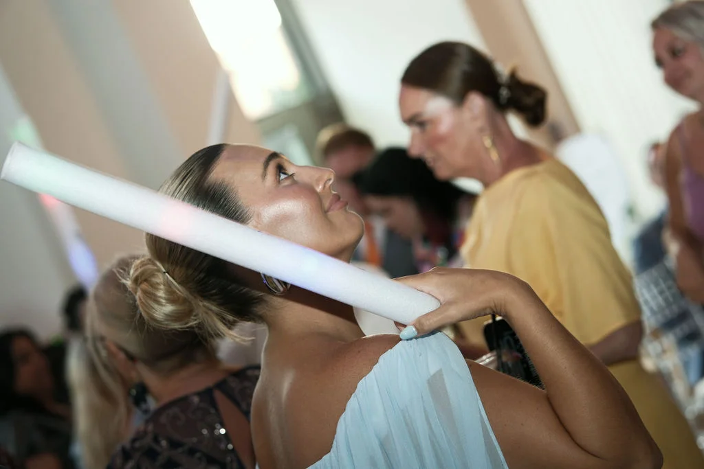 A guest at a wedding reception looks up while holding a light-up foam baton over her shoulder amidst a crowd of mingling guests.
