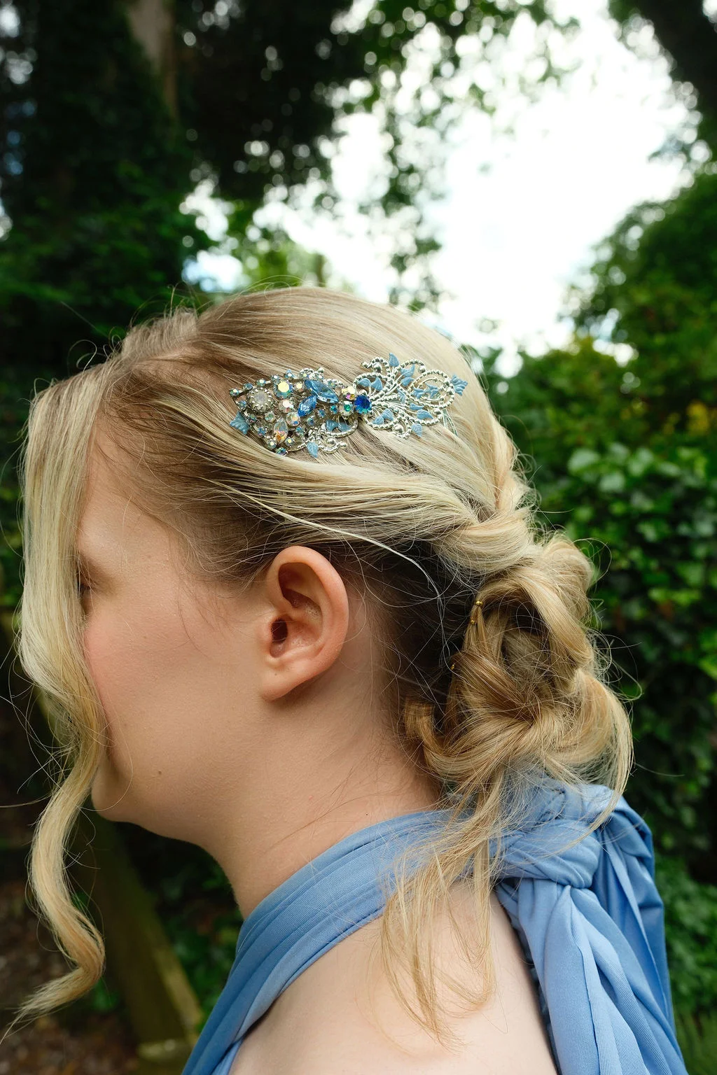 A blonde bride wearing an intricate crystal hair comb and a blue dress, viewed from the side outdoors against a lush green background.