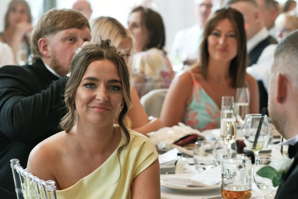 A guest smiles towards the camera while seated at a wedding reception table surrounded by other diners and drinks.