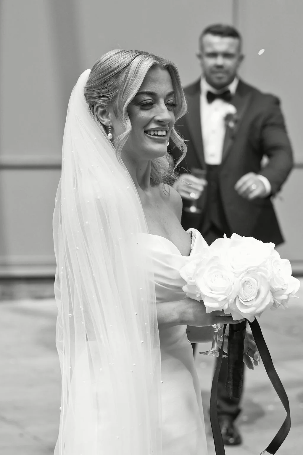 A smiling bride wearing a strapless gown and pearl veil holds a bouquet of white roses while the groom watches lovingly in the background.