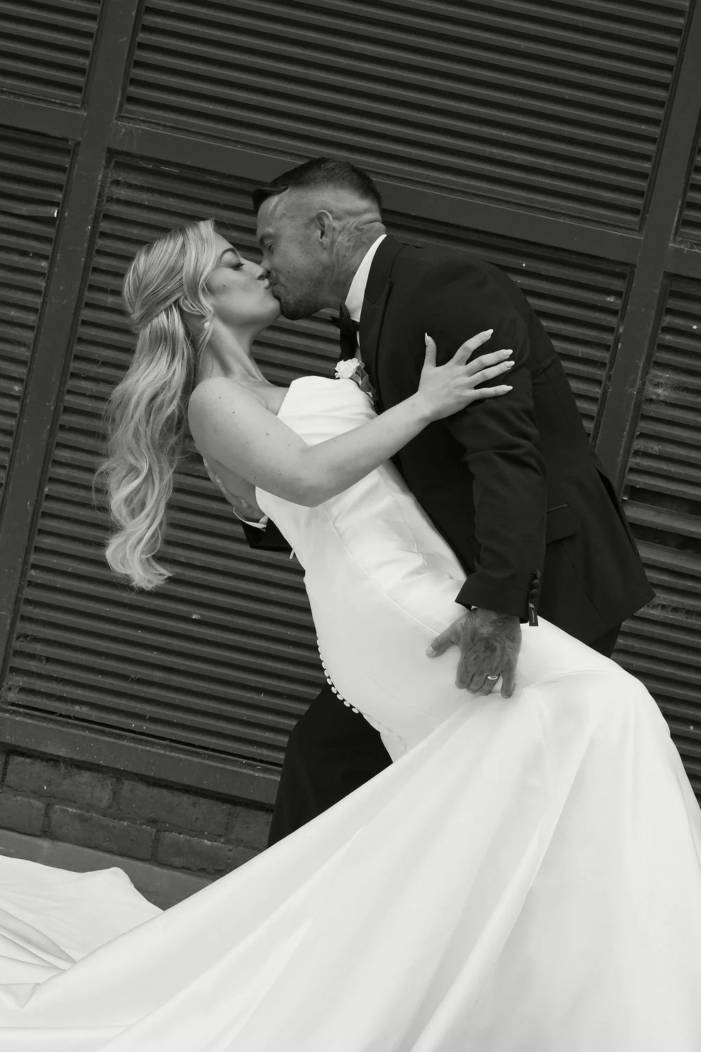 A newlywed couple shares a dramatic dip kiss in front of a slatted dark wall, with the bride wearing a sleek strapless satin gown and the groom in a classic black tuxedo.