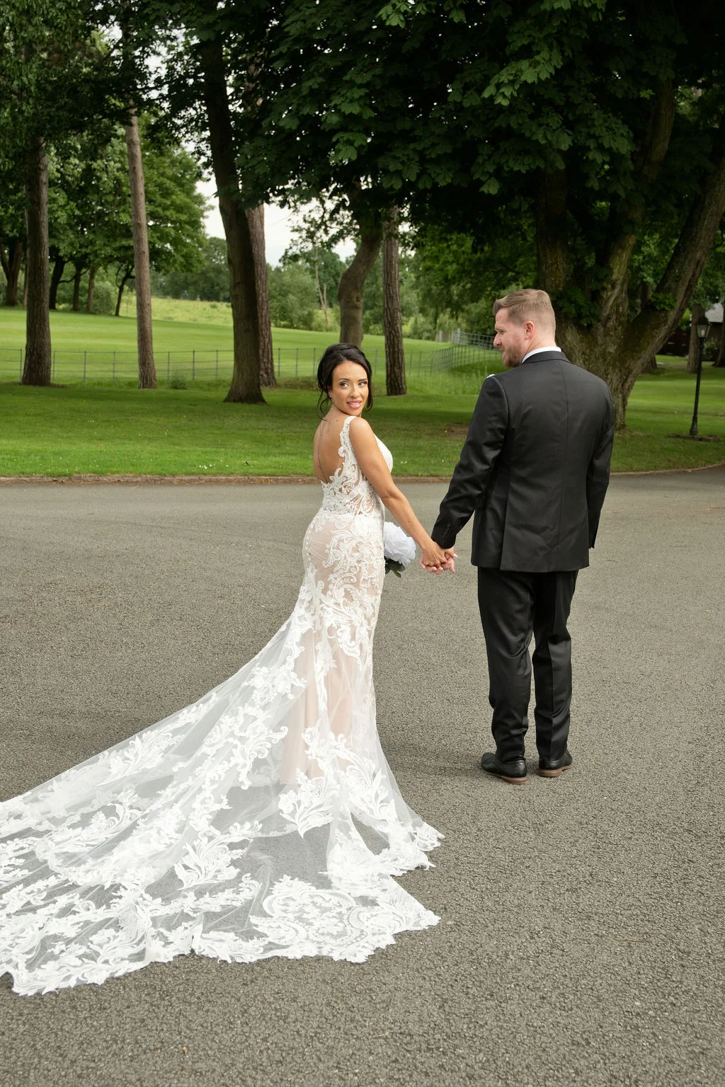 A newlywed couple strolls hand in hand along a tarmac path, with the bride glancing back over her shoulder to reveal her stunning lace gown featuring a dramatic, cascading train.