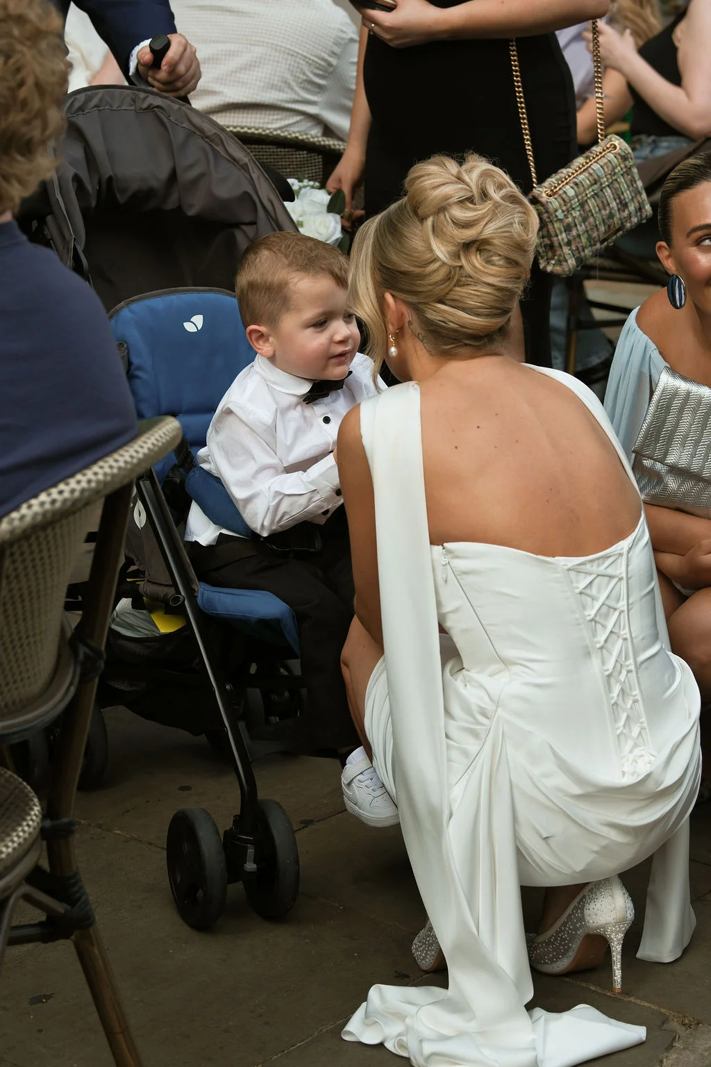 A bride in an elegant white gown with a lace-up back crouches down to chat with a young page boy sitting in a pushchair among wedding guests.