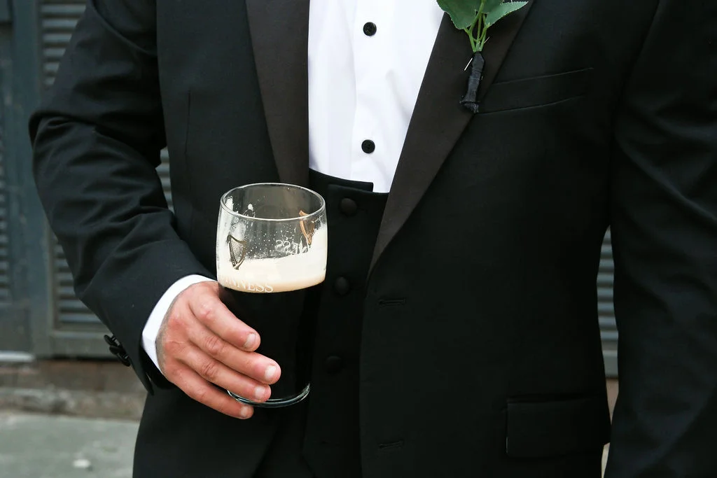 A groom dressed in a sharp black tuxedo with a buttonhole pinned to his lapel holds a pint glass of dark stout.