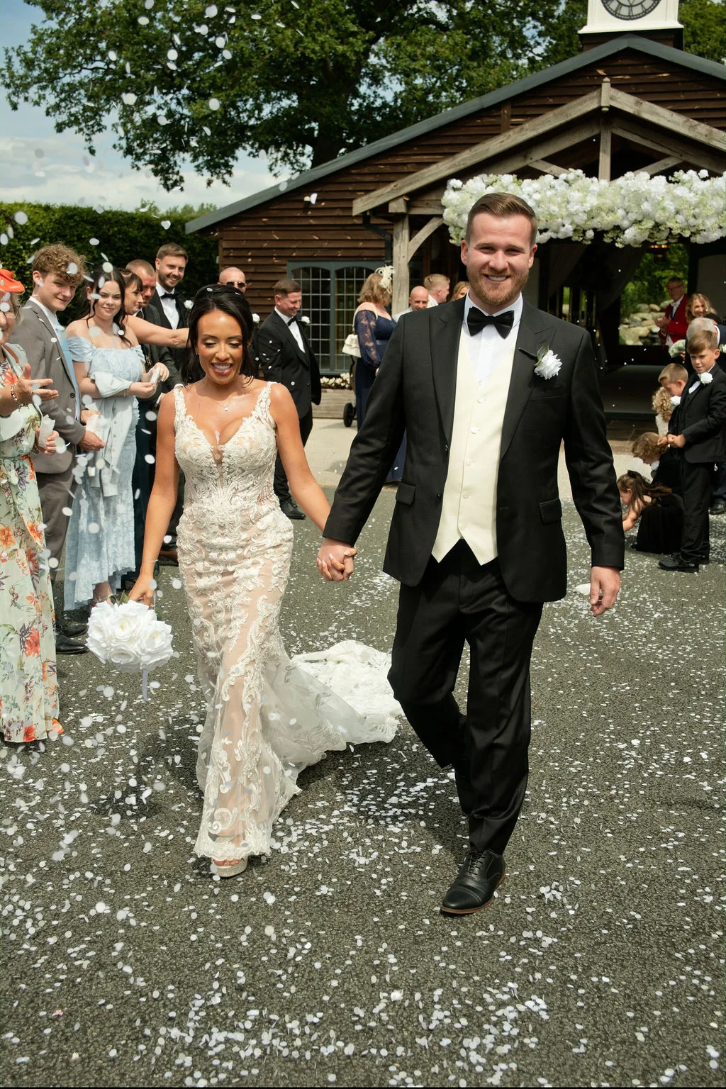 Newlyweds walk hand in hand down the aisle smiling as guests shower them with white confetti outside a rustic barn venue.