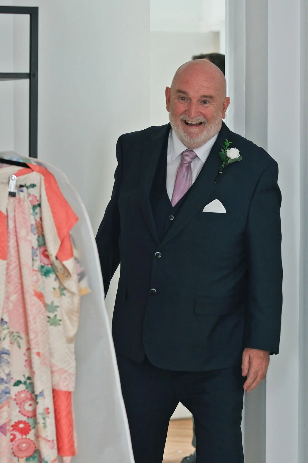 A smiling father of the bride wearing a navy three-piece suit and a white floral buttonhole reacts with joy during wedding preparations.