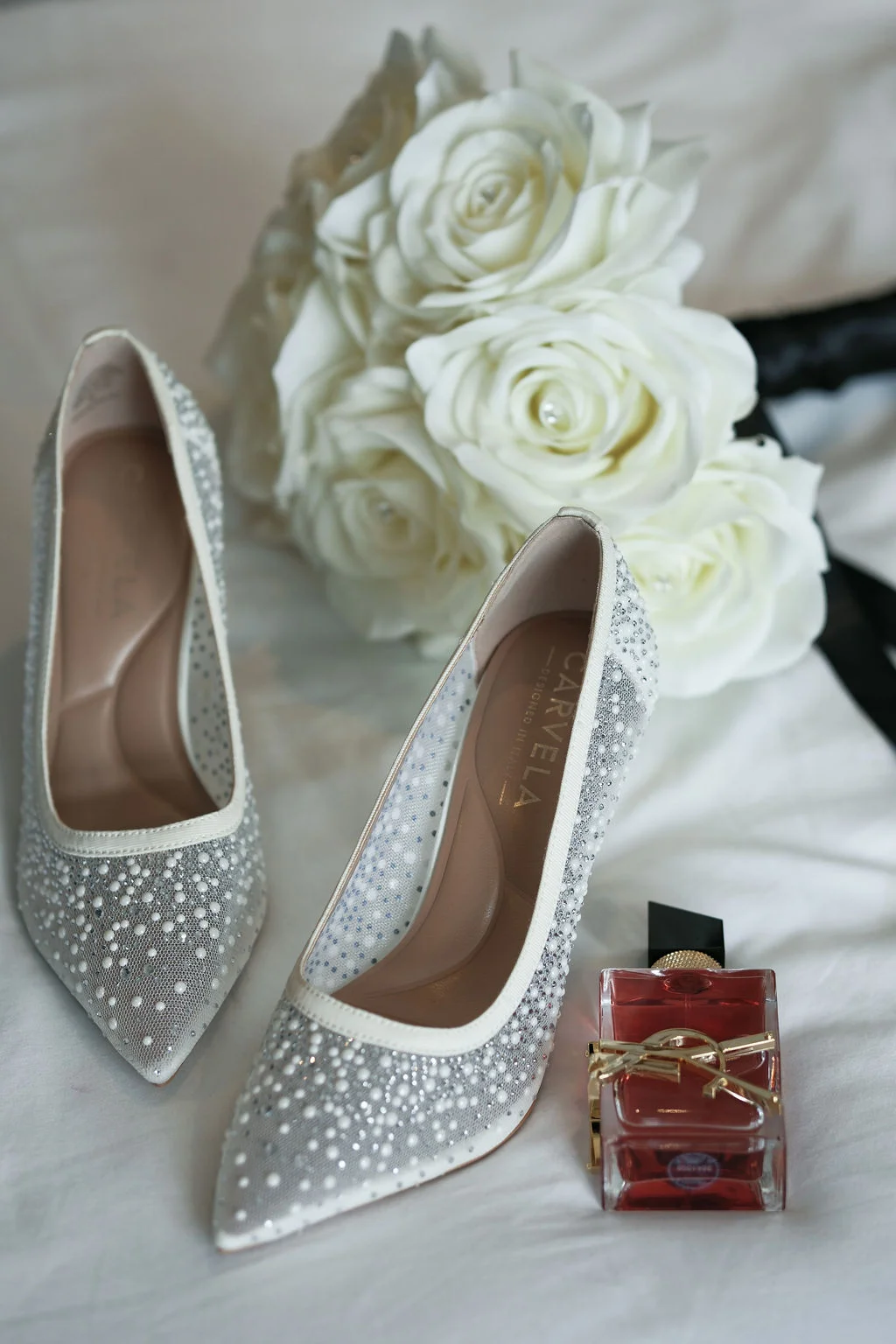 An elegant bridal flat lay features a pair of crystal and pearl-embellished heels alongside a bouquet of white roses and a bottle of perfume.
