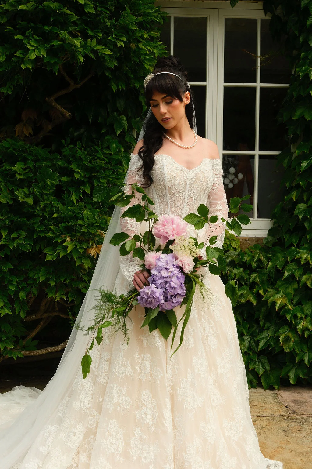 A stunning bride poses in front of an ivy-covered wall, holding a lush cascading bouquet filled with pink and purple blooms while wearing an off-the-shoulder lace gown.