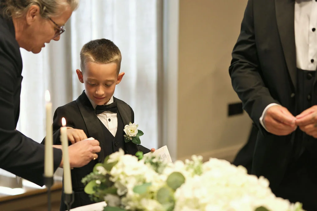 A young page boy in a smart black tuxedo and bow tie stands near a candlelit table decorated with white floral arrangements during a wedding ceremony.