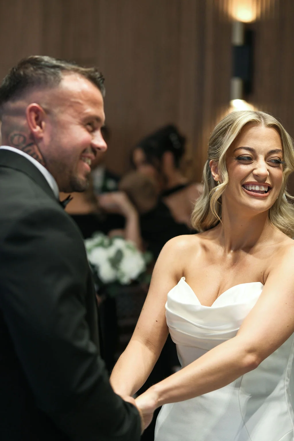 A joyful newlywed couple holding hands and laughing during their wedding ceremony inside a contemporary venue.