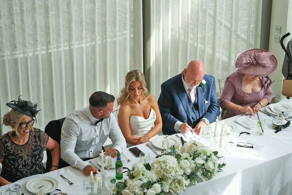 A bridal party and family members are seated at a top table during a wedding breakfast, with guests gathered around and floral arrangements decorating the setting.