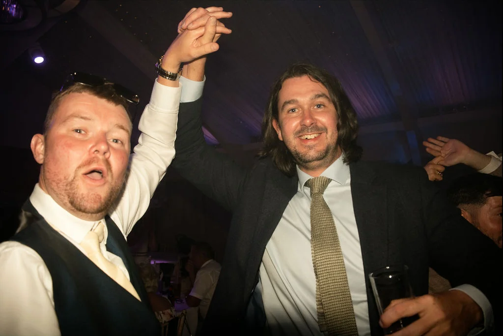 Two smiling wedding guests dance together on the evening dance floor with their hands raised in the air.