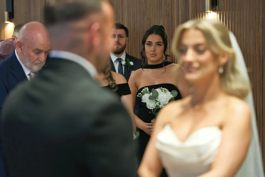A bride in a strapless white gown and groom stand during their wedding ceremony while bridesmaids in black dresses look on.