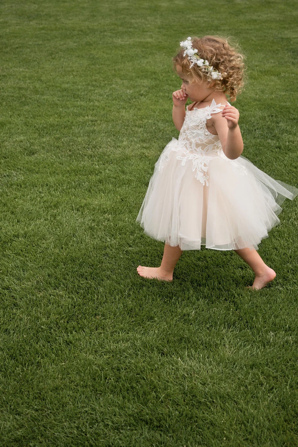 A barefoot flower girl with curly hair wearing a tulle dress and a floral crown walks across a green lawn.