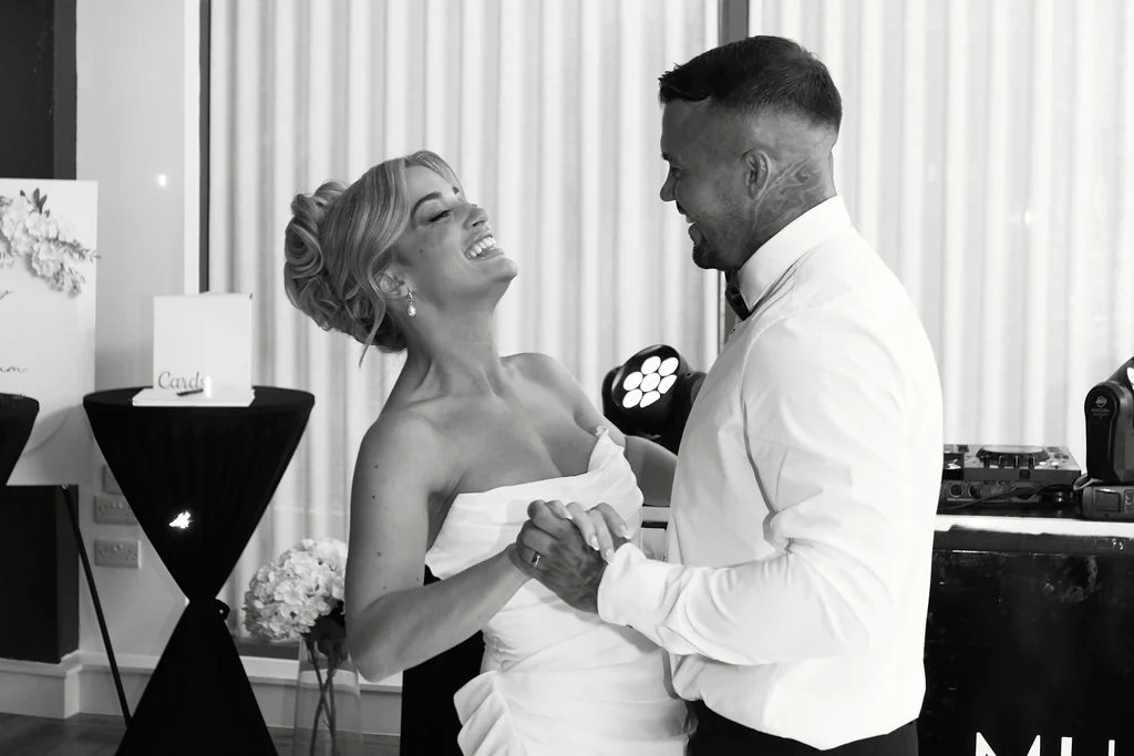 A newlywed couple shares a joyful laugh during their first dance, with the bride leaning back in her strapless white dress while holding hands with the groom.