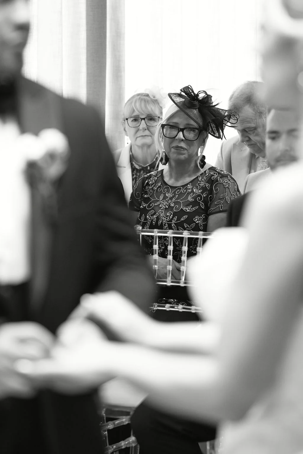 An emotional wedding guest wearing a fascinator watches attentively as the couple exchanges rings during the ceremony in soft focus.
