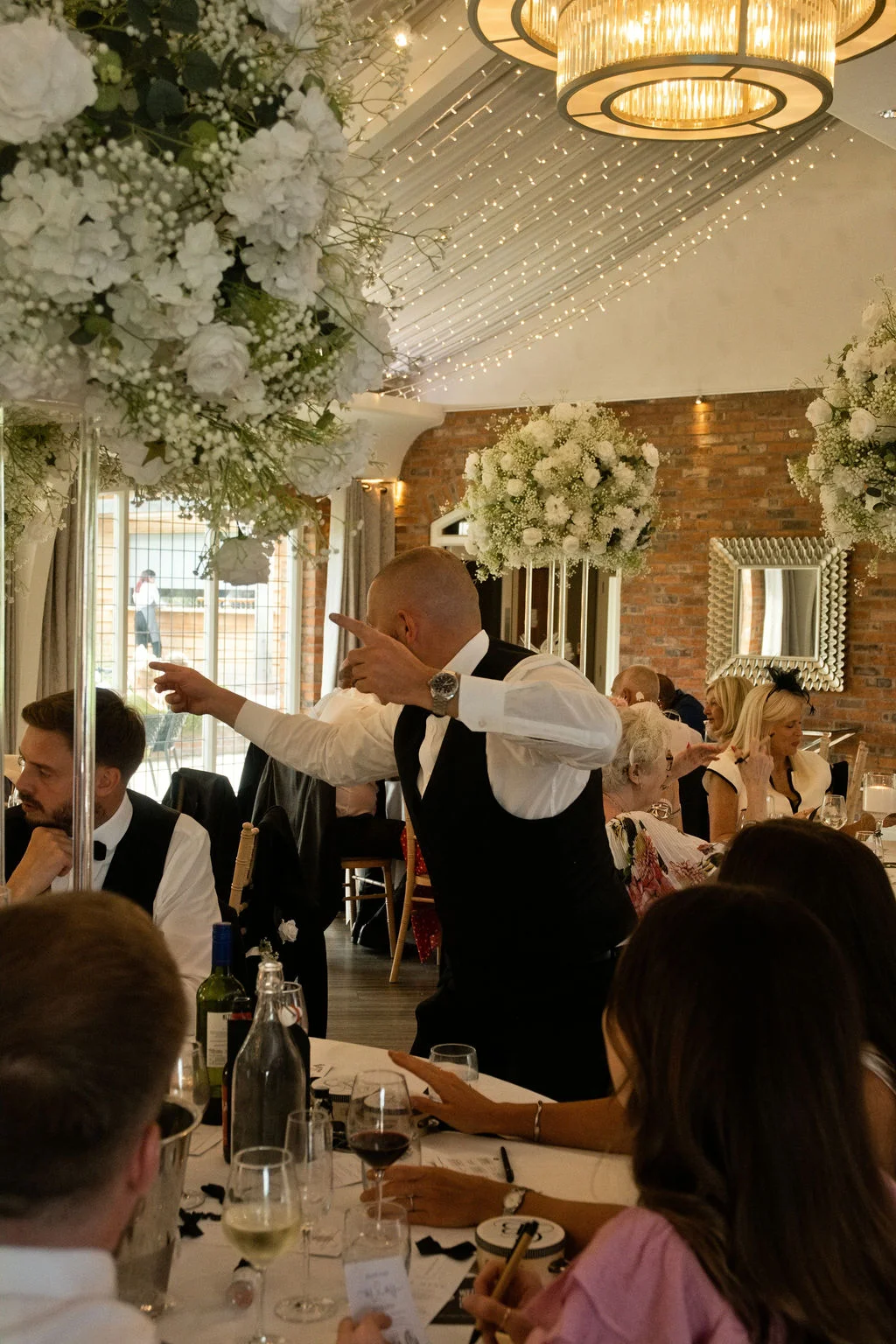 A lively wedding reception features guests seated at a long banquet table under a canopy of fairy lights, with a guest standing and gesturing animatedly during speeches.