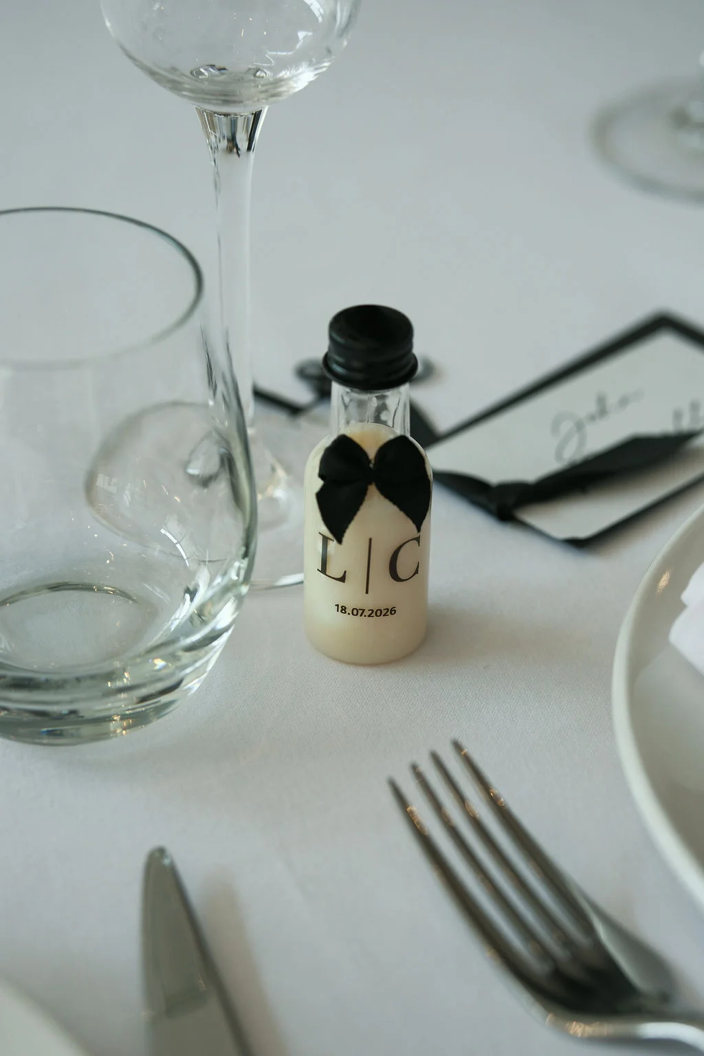A close-up of a wedding place setting featuring a monochrome name card and a miniature favour bottle decorated with a black bow and monogram.