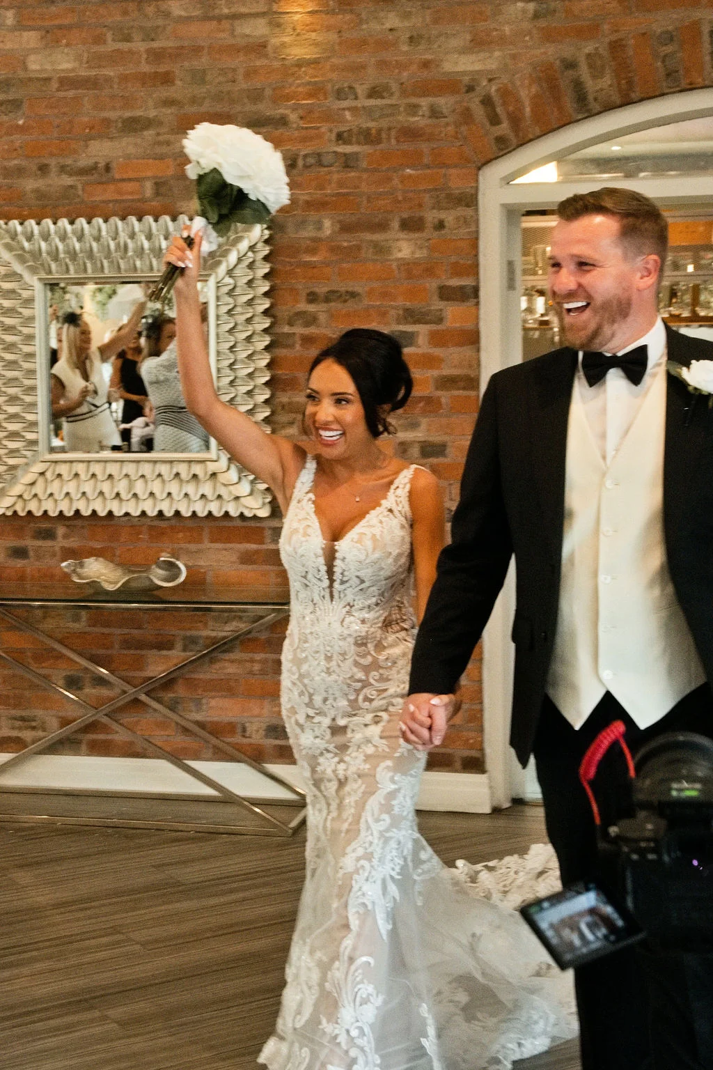 A newlywed couple excitedly enters their wedding reception, with the smiling bride holding her bouquet high in the air while holding hands with the groom.