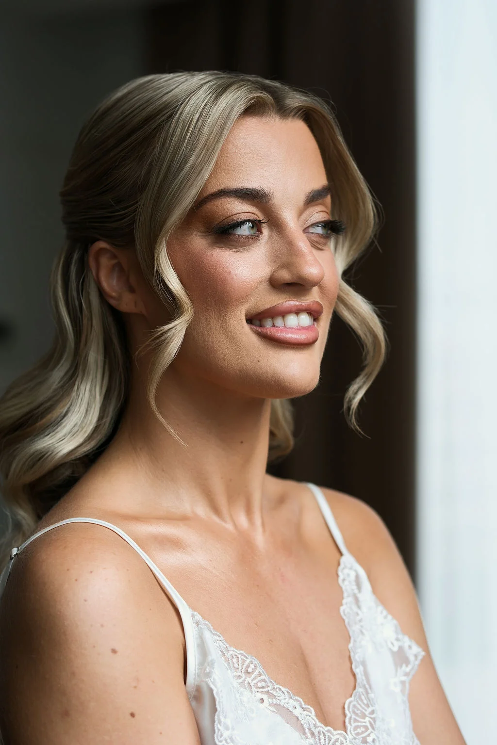 A stunning portrait of a smiling bride getting ready in soft window light, showcasing her half-up blonde hairstyle and delicate lace robe.