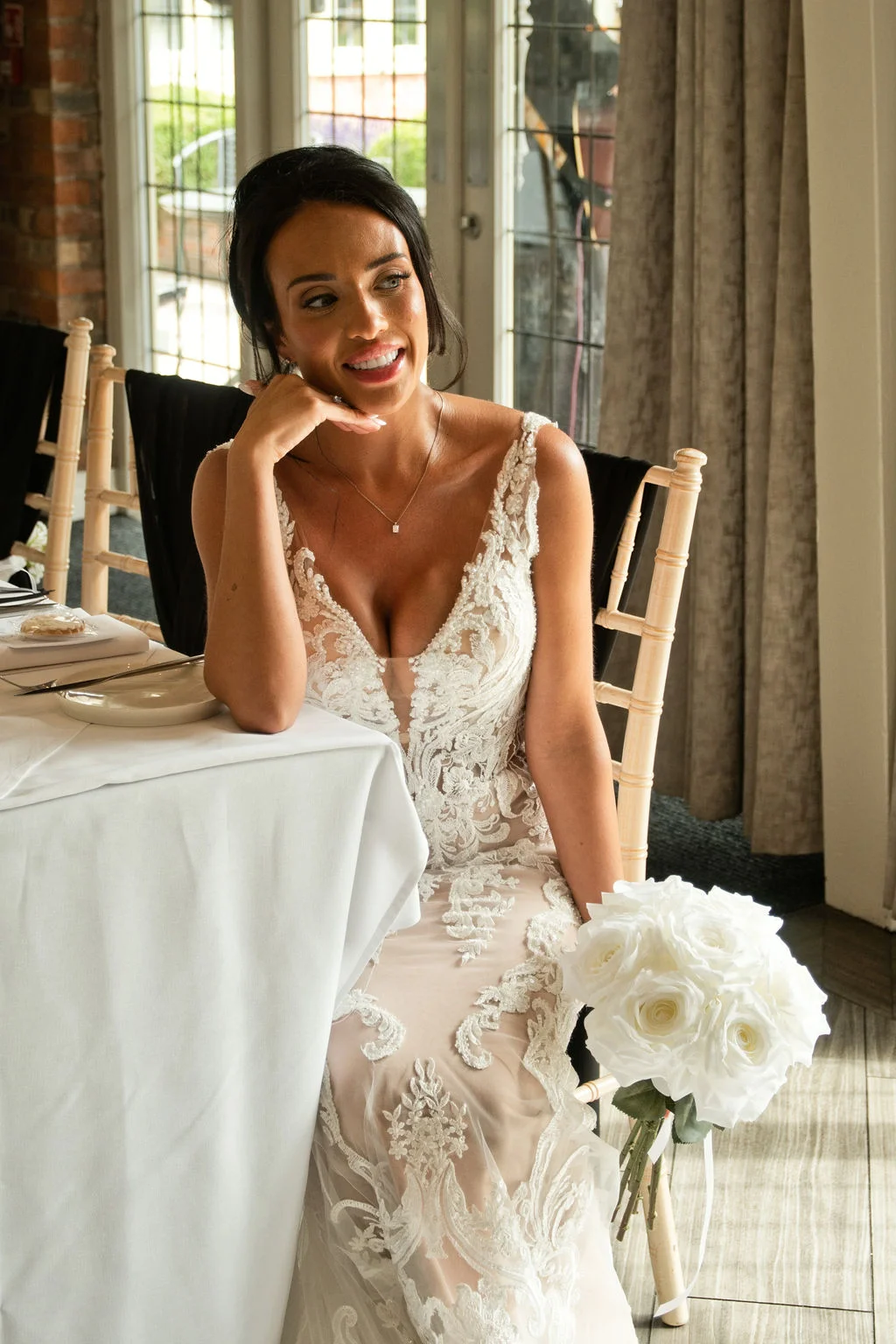 A smiling bride wearing a lace illusion wedding dress sits at a reception table holding a round white rose bouquet.
