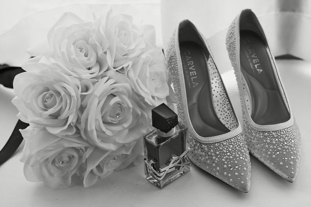 A sophisticated monochrome flat lay features a sparkling bridal shoe pair alongside a lush white rose bouquet and a designer perfume bottle.