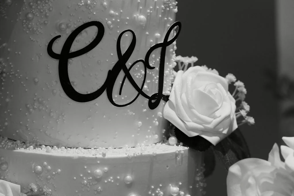 A detailed close-up shot of a white wedding cake adorned with pearls, a black monogram cake topper, and white roses in a monochrome finish.