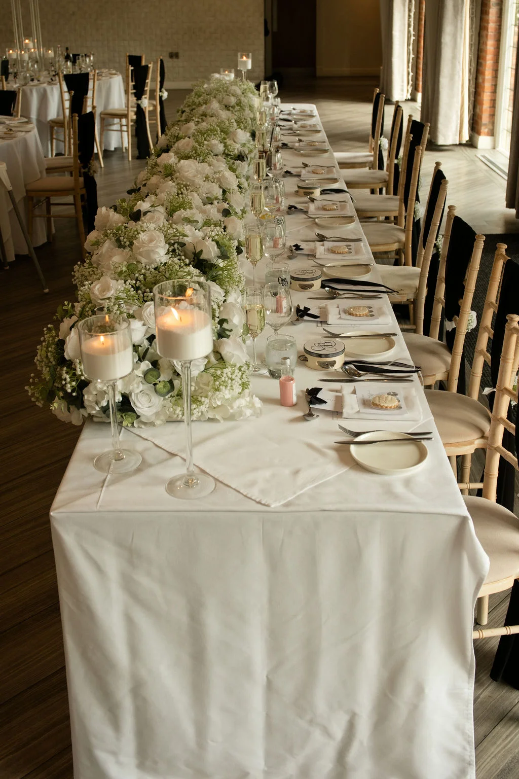 An elegant wedding reception room features a long banquet table draped in white linen and adorned with an abundant floral runner of white roses and gypsophila, tall pillar candles in glass holders, and meticulously arranged place settings.