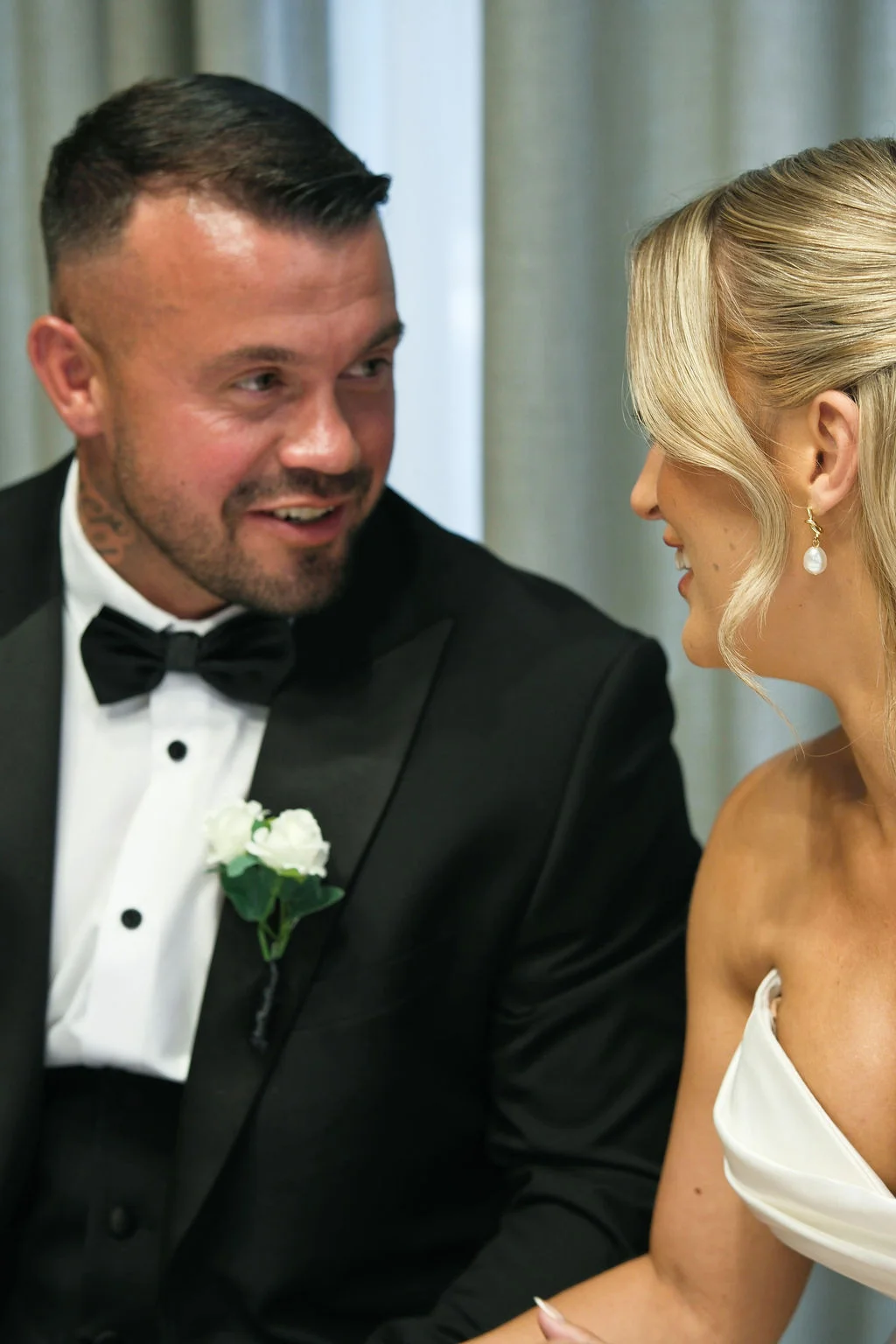 A smiling groom in a black tuxedo and bow tie gazes lovingly at his bride, who wears elegant pearl drop earrings and a strapless white wedding gown.