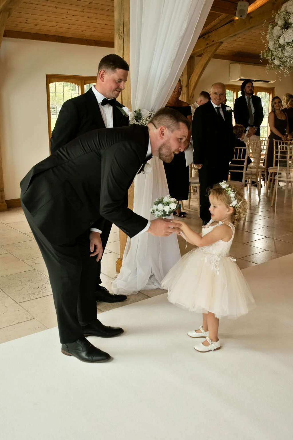 A groom in a black tuxedo leans down to gently greet a young flower girl holding a tiny bouquet during a bright indoor ceremony.