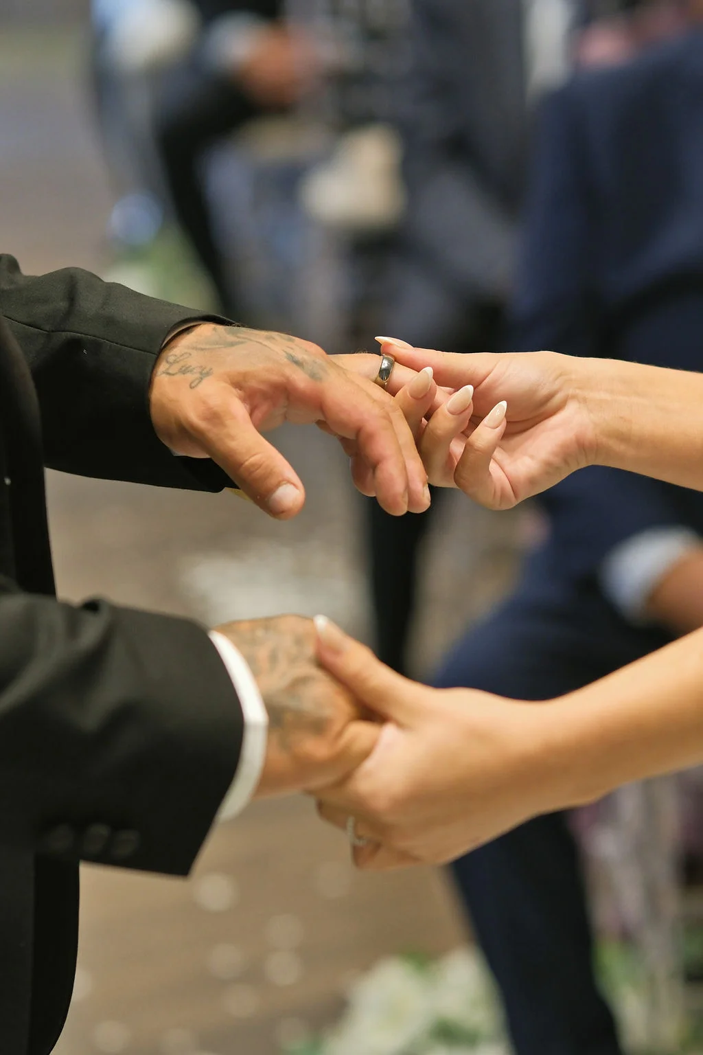 Close-up shot capturing a couple exchanging wedding rings during the ceremony, with visible tattoos on the groom's hand and delicate manicured nails on the bride.