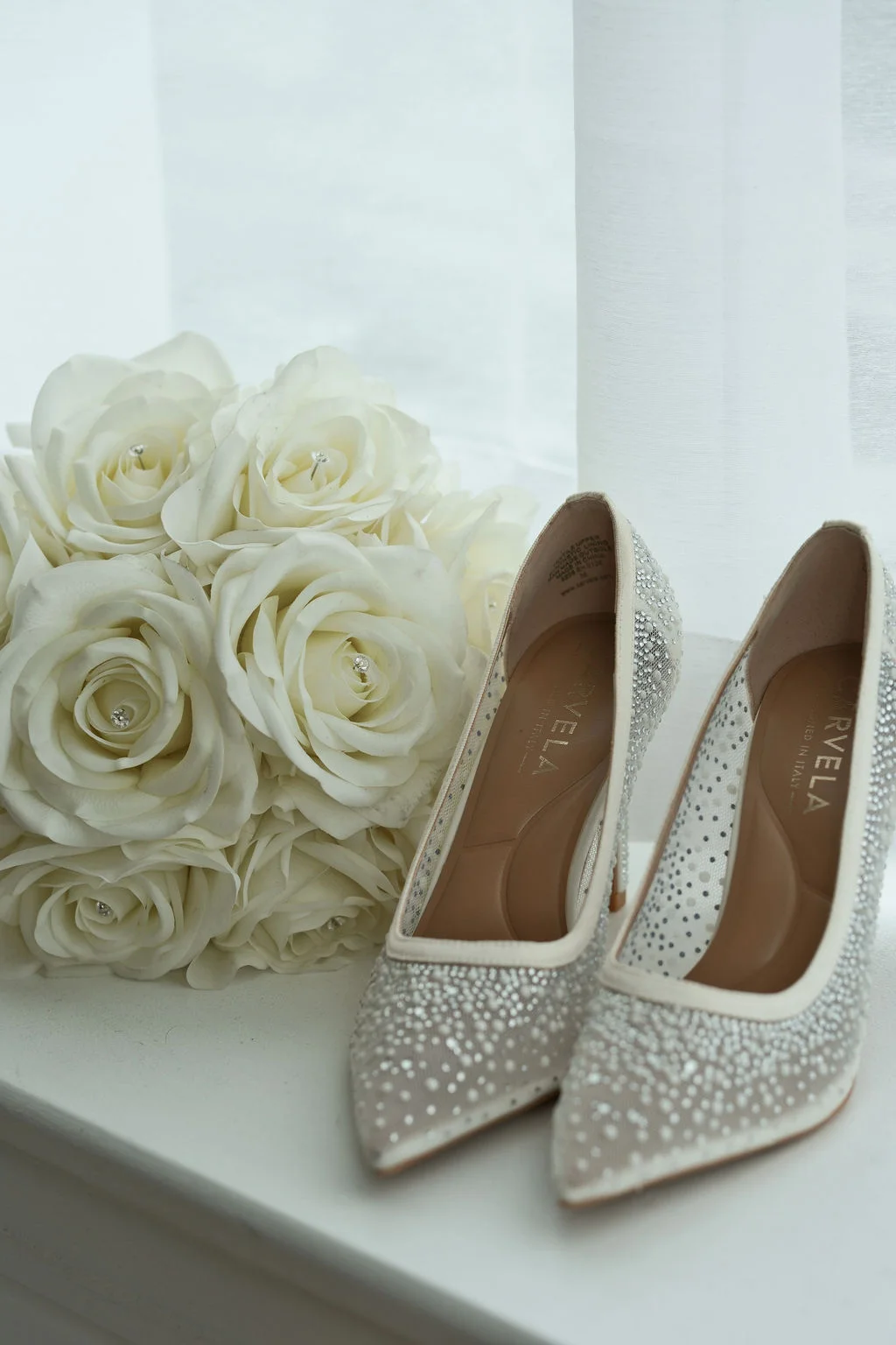 A close-up flat lay showcases a pair of sparkly bridal heels resting beside a lush arrangement of white roses near a bright window.