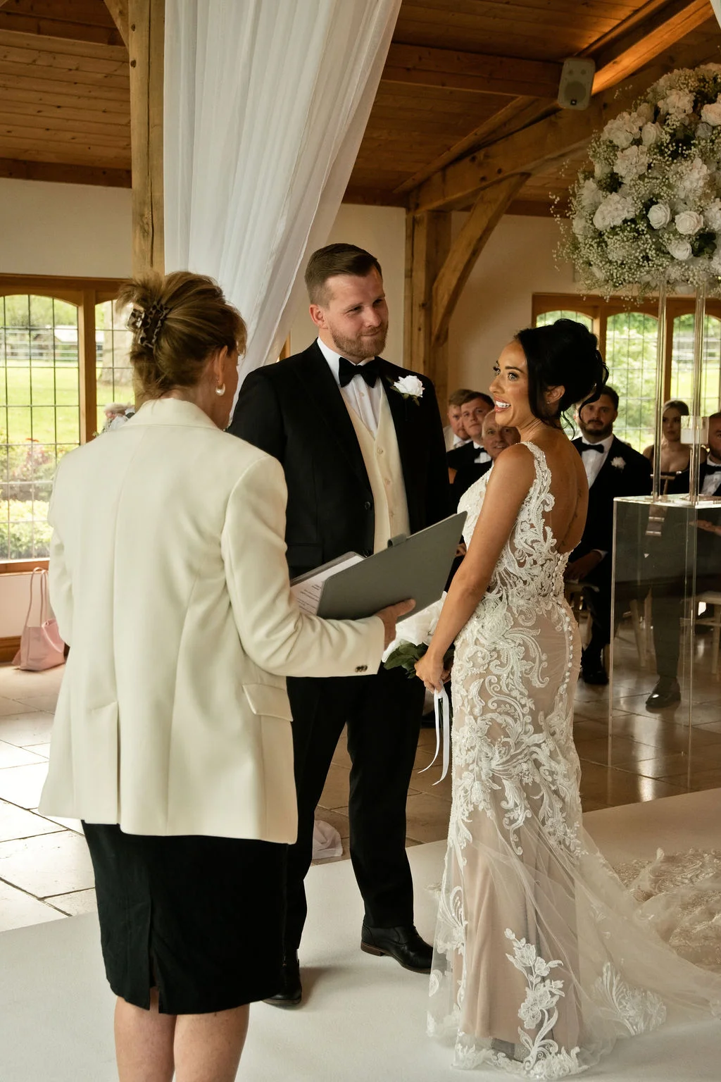 A bride and groom exchange vows during an indoor civil ceremony inside a light-filled barn venue with wooden beams and white floral arrangements.